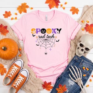 May include: A light pink t-shirt featuring the text SPOOKY rad tech in black, orange, and purple. The design includes a spider web, a spider, bats, and small star icons. The shirt is displayed with a pumpkin, orange sneakers, and a skull.