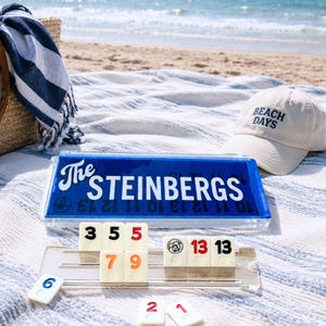 May include: A blue and clear acrylic game case with the white text The Steinbergs printed on the front. Below the case are several off-white rectangular game tiles with black, red, and orange numbers, resting on a clear plastic tile rack.