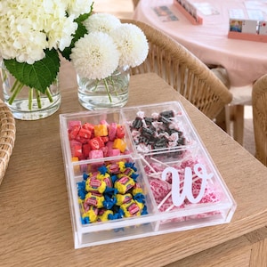 May include: A clear acrylic four-compartment candy organizer box filled with assorted colorful wrapped candies. The bottom right section features a white cursive letter W decal on the lid. The container is square with transparent walls.
