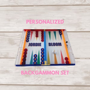 May include: A personalized backgammon set featuring a clear acrylic board with orange and blue borders. The board displays the names JORDIE BLOOM in blue text, with red and blue circular checkers, dice, and colorful triangular point markers.