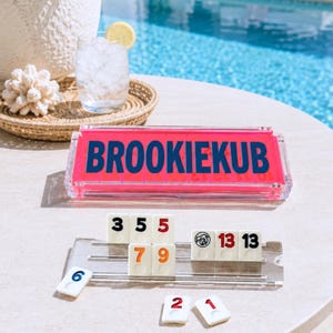 May include: A clear acrylic tile rack for a rummy-style game featuring the name BROOKIEKUB in bold blue letters on a bright pink background. The set includes white game tiles with black, red, orange, and blue numbers and a joker icon.