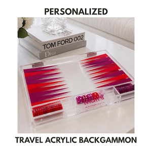 May include: A clear acrylic backgammon set featuring a board with alternating red and purple triangular points. The set includes red and purple game pieces, dice, and a doubling cube. The text "PERSONALIZED" and "TRAVEL ACRYLIC BACKGAMMON" is visible.