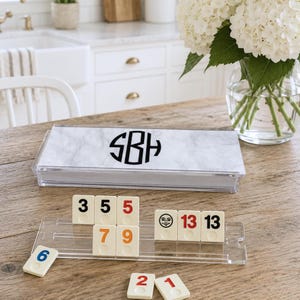 May include: A rectangular acrylic box with a white marble pattern and a black monogram featuring the letters SBH. Below the box is a clear acrylic tile rack holding cream-colored game tiles with numbers in black, red, orange, and blue, plus a moon icon.