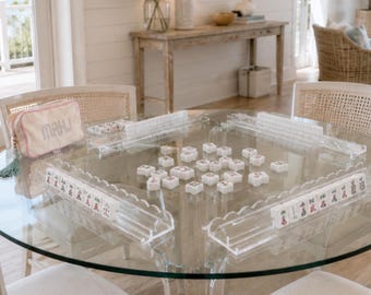 Op de afbeelding: Een mahjongspel van helder acryl met witte tegels voorzien van rode en zwarte markeringen, opgesteld op een glazen tafel met vier transparante rekjes. Een crèmekleurig tasje met rits en het woord MAHJ in wit borduurwerk ligt op een stoel.