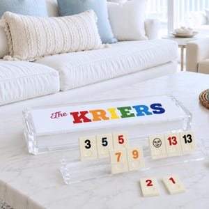 May include: A clear acrylic game set featuring a rectangular box with the text The KRIERS printed in multicolored letters. The set includes a matching clear acrylic tile rack holding cream-colored tiles with numbers and a moon face icon.