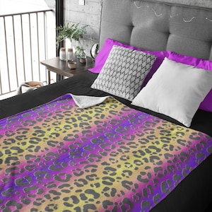 May include: A colorful blanket with a leopard print design. The blanket features a gradient of yellow, pink, and purple hues, with dark gray leopard spots. The blanket is placed on a bed with black sheets and decorative pillows.