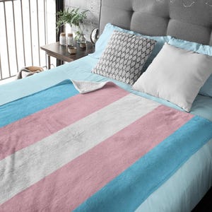 Pode incluir: Uma manta macia e felpuda com as cores da bandeira do orgulho transgénero: azul claro, rosa e branco. A manta está dobrada e exposta numa cama com almofadas.