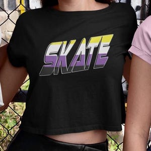 May include: Black cropped t-shirt with the word "SKATE" in yellow, purple, and black lettering. The lettering is in a bold, 3D style.