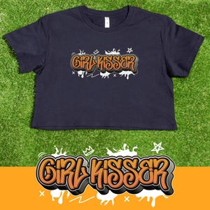 May include: A navy blue crop top featuring the text "GIRL KISSER" in orange and grey graffiti lettering. The design incorporates crowns, stars, and dripping paint effects. The top is set against a green backdrop.