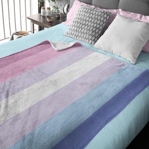 Könnte beinhalten: Eine weiche, gestreifte Wolldecke mit rosa, weißen, lavendelfarbenen, hellblauen und dunkelblauen Streifen. Die Decke liegt auf einem Bett mit einem hellblauen Laken.