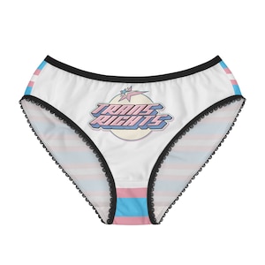 Puede incluir: Bóxer blanco con cintura negra y ribete de encaje. La ropa interior tiene un patrón de bandera del orgullo transgénero azul y rosa y un gráfico que dice "Trans Rights" en letras rosas y azules.