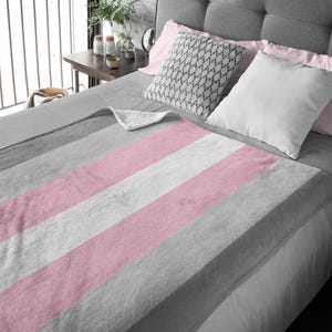 Könnte beinhalten: Eine weiche, plüschige Decke mit abwechselnden rosa und grauen Streifen. Die Decke ist gefaltet und über ein Bett mit Kissen gelegt.