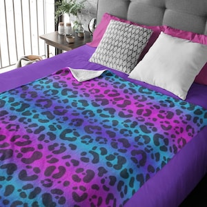 Pode incluir: Uma manta de cor azul e roxo degradê com estampa de leopardo preto. A manta está dobrada e colocada em uma cama roxa.