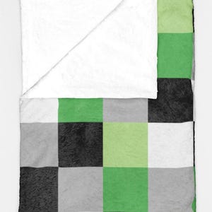 Pode incluir: Uma manta macia e felpuda com um padrão xadrez em tons de verde, preto, cinza e branco. O canto superior está dobrado, revelando um tecido branco liso. A manta foi projetada para conforto e calor.