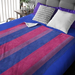 Pode incluir: Uma manta de lã azul e rosa em listras sobre uma cama azul com almofadas de padrões brancos e cinzentos.