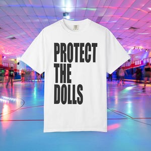 Puede incluir: Camiseta blanca con el texto "PROTECT THE DOLLS" en letras negras y gruesas. La camiseta se muestra en una pista de patinaje sobre ruedas con gente patinando al fondo. Una prenda de vestir informal.