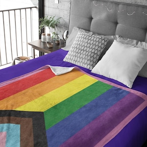 Könnte beinhalten: Eine lilafarbene Fleece-Decke mit einem Regenbogen- und braunen Streifenmuster. Die Decke ist auf einem Bett mit weißen und grauen Kissen gefaltet.