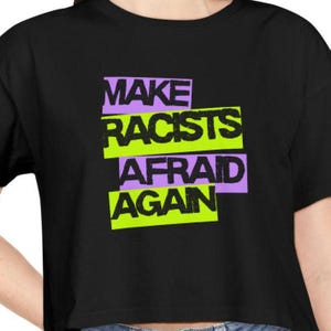 Puede incluir: Top corto negro con el texto "MAKE RACISTS AFRAID AGAN" en letras de bloque verde neón y morado. Una prenda de vestir informal.