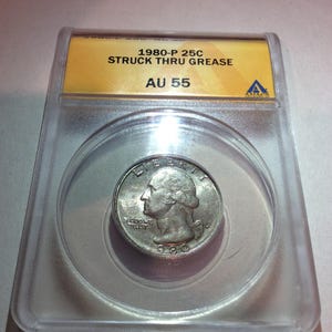 May include: A 1980-P 25-cent coin, graded AU 55, encased in a clear plastic holder. The coin features a profile of George Washington and the words "LIBERTY" and "IN GOD WE TRUST". The holder has a yellow label with black text.