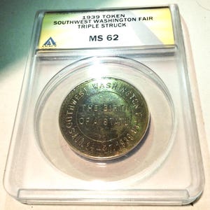 May include: A vintage coin from the 1939 Southwest Washington Fair, triple struck, encased in a clear protective holder. The coin features text that reads "The Birth of a State" and "Southwest Washington Fair". The holder is labeled "MS 62".