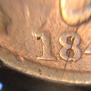 May include: Close-up of a copper coin fragment with the numerals "18" and part of "84" visible. The coin's surface shows signs of wear and age, with visible cracks and discoloration. The coin's details are partially obscured.