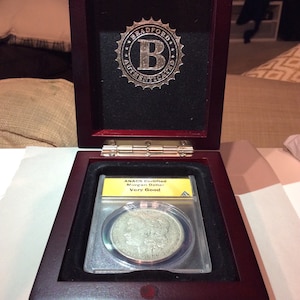 May include: A wooden display case with a silver coin inside. The coin is encased in a clear plastic holder with a yellow label that reads "ANACS Certified Morgan Dollar Very Good". The case has a "Bradford Authenticated" emblem.