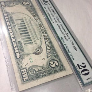 May include: A five-dollar bill from 1988, encased in a protective holder. The bill shows an offset printing error and is graded as Very Fine. The holder has text indicating the Federal Reserve Note and the serial number.