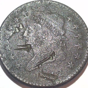 May include: A detailed close-up of an antique coin with a profile of a person, possibly a historical figure. The coin is a dark, aged silver color, with visible wear and tear. Stars and other design elements are also visible.