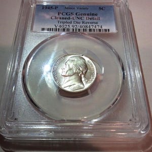 May include: A 1945-P Jefferson nickel encapsulated in a clear plastic PCGS slab. The label reads 1945-P Minor Variety 5C, PCGS Genuine Cleaned-UNC Detail, Tripled Die Reverse, and V4025.92/40847474. The silver-toned coin shows a profile of Thomas Jefferson.