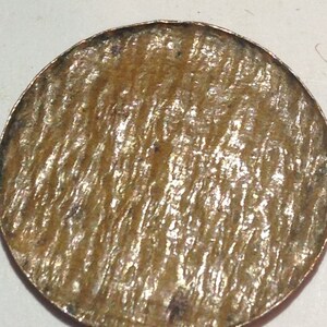 May include: A close-up of a circular, metallic object with a textured surface. The object has a bronze-like appearance with a slightly uneven edge. The surface has a pattern of parallel lines.