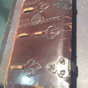 May include: A vintage, leather-bound book with the title "The Autobiography" embossed on the cover. The dark brown cover features decorative leaf-shaped accents and metal clasps. The book's aged appearance suggests it is an antique or a replica.