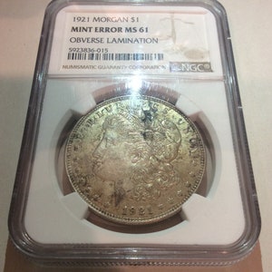 May include: A 1921 Morgan silver dollar coin, graded MS 61 with an obverse lamination error, encased in a protective holder. The coin features a profile of a woman and the text "LIBERTY". The holder includes the text "NUMISMATIC GUARANTY CORPORATION".
