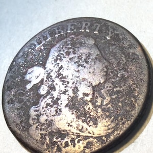 May include: A silver coin with a portrait of a woman and the word "LIBERTY" at the top. The coin has a worn, aged appearance, with visible signs of wear and oxidation. The coin's design includes a profile and the date.