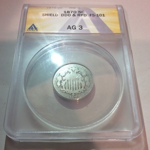 May include: A silver 1870 Shield nickel coin, encapsulated in a clear plastic holder. The coin features a shield design with the year 1870 visible. The holder has a label with text including "1870 5C SHIELD DDO & RPD FS-101" and "AG 3".