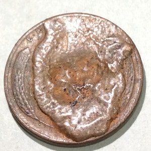 May include: A close-up of a copper-colored coin with significant wear and corrosion. The coin's surface is heavily textured, with raised areas and visible damage. The design is partially visible, with what appears to be a profile of a person.