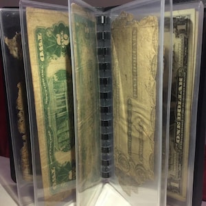 May include: A clear plastic currency holder with several old U.S. dollar bills visible. The bills are in various states of wear, with denominations including one and five dollar bills. The holder is bound with a black spine.