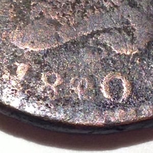 May include: Close-up of a weathered coin with the year 1890 visible. The coin has a textured surface with a dark, aged appearance. The numbers are slightly raised and worn.