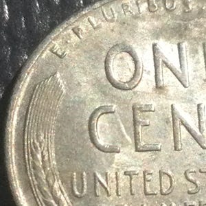 May include: Close-up of a well-worn United States one-cent coin. The coin features the words "ONE CENT" and "UNITED STATES OF AMERICA". A wheat stalk design is visible on the left side of the coin.