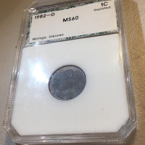 May include: A 1982-D MS60 unplated one-cent coin encased in a clear plastic holder. The coin is centered on a white background with the text "1C Unplated" and "1982-D MS60" visible. The mintage is unknown.