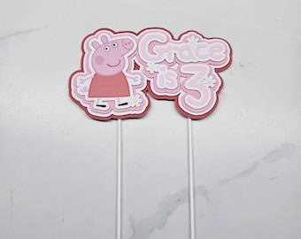 May include: A layered paper cake topper featuring a pink cartoon pig character and the text "Grace is 3" in a light pink font with white outlines. The design includes small white daisy accents and is supported by two thin white sticks.