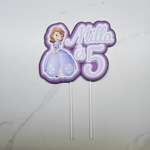 May include: A layered paper cake topper featuring a cartoon princess in a purple dress with the text Milla is 5. The design uses shades of purple and white, with two white sticks attached to the bottom for insertion into a cake or dessert item.