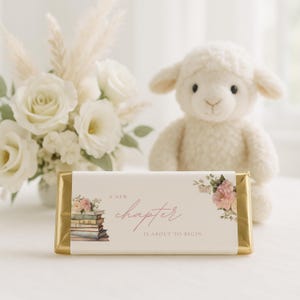 Puede incluir: Una barra de chocolate envuelta con el texto "A New Chapter Is About To Begin" e ilustraciones florales. Un cordero de peluche y un ramo de flores blancas están en el fondo. La barra tiene un envoltorio de papel dorado.