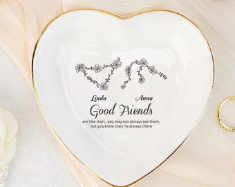 Op de afbeelding: Een hartvormige witte keramische sieradenschaal met een gouden rand. De schaal bevat de woorden "Linda" en "Anna" boven de zin "Good Friends" en een bloemenontwerp. De tekst "are like stars, you may not always see them, but you know they're always there" is ook opgenomen.