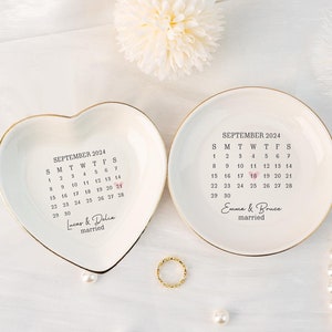 Può includere: Due porta anelli in ceramica bianca con bordo dorato. Un piatto è a forma di cuore e l'altro è rotondo. Entrambi i piatti presentano un calendario per settembre 2024 con la data 18 evidenziata. Il piatto a forma di cuore ha il testo "Lucas & Delia sposati" e il piatto rotondo ha il testo "Emma & Bruce sposati".