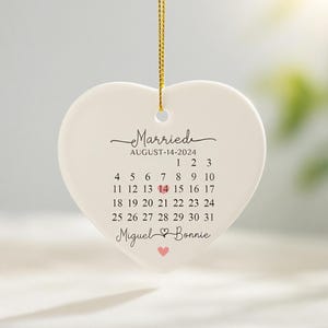 May include: A white heart-shaped ceramic ornament hanging from a gold string. It features the text Married, August-14-2024, a calendar for August with the 14th highlighted by a pink heart, and the names Miguel and Bonnie with a small pink heart.