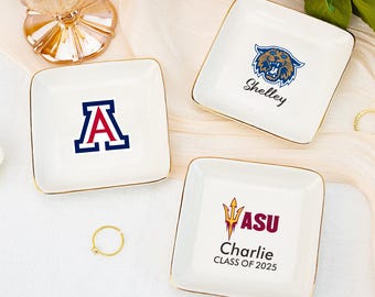 Puede incluir: Tres platos cuadrados de cerámica blanca con bordes dorados. Cada plato presenta un logotipo y un nombre universitario diferente. Uno tiene un logotipo de tigre azul y dorado con el nombre "Shelley". Otro tiene el logotipo "ASU" y "Charlie CLASS OF 2025". El tercero tiene una "A" azul y roja.
