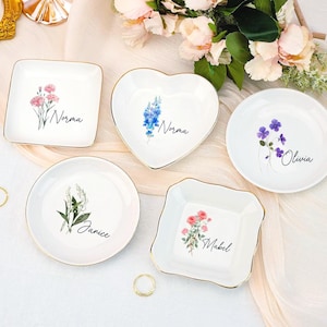 Puede incluir: Cinco platos de cerámica blanca para baratijas con bordes dorados, cada uno con un diseño floral y un nombre: Norma, Olivia, Janice y Mabel. Un plato tiene forma de corazón. Los platos están dispuestos sobre una superficie blanca con flores.