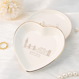 以下が含まれることがあります： 金色の縁取りが施された白いハート型のセラミック製の小物入れ。ロンドンのスカイラインが線画で描かれており、ビッグベン、タワーブリッジ、国会議事堂などが描かれている。スカイラインの下には「Richard & Farisa 10.10.2024」という文字が印刷されている。