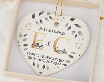 Pode incluir: Um ornamento de cerâmica em forma de coração numa caixa de presente bege, com um design de coroa de folhas verdes. O texto diz: JUST MARRIED, E & C, Elliot & Cecilia, HAPPILY EVER AFTER, 20TH AUGUST 2024. Um cordão dourado está preso ao topo.