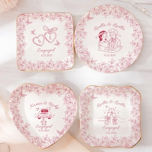 Pode incluir: Quatro pratos de cerâmica brancos para noivado com desenhos florais rosa e bordas douradas. Cada prato tem uma forma diferente e apresenta a palavra "Engaged" e uma data. Os pratos têm ilustrações de corações, um casal, um carro e taças de champanhe.
